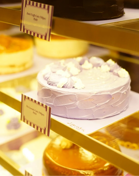 A bakery display case showing a frosted lavender cake with piped cream decorations on the middle shelf, alongside other assorted cakes and small product labels.