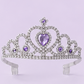 A product lineup graphic showing lavender and silver party accessories on a light lavender background, including a gold and lavender crown, lavender snow spray cans, a silver tiara, a lavender party popper, a lavender spiral candle, and two silver sparkles.