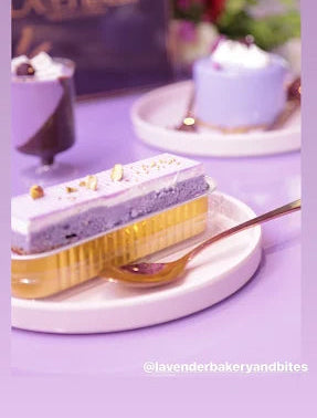 A close-up of a lavender and purple layered cake slice on a white plate with a gold spoon, set against a purple background with other desserts and a blurred sign in the distance.